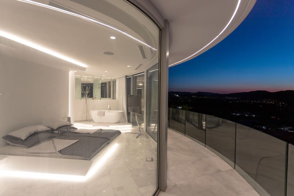 Chambre minimaliste avec sol en marbre, éclairage LED intégré sous le lit, baignoire îlot et terrasse courbe offrant une vue panoramique sur la vallée au crépuscule.