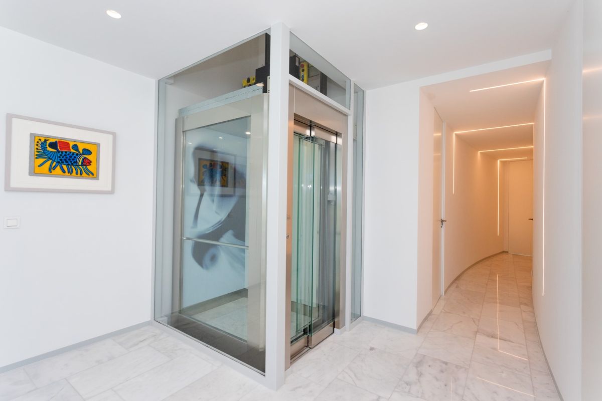 Couloir moderne avec ascenseur vitré aux finitions en acier, sol en marbre et un dégagement éclairé par des bandes LED linéaires intégrées.