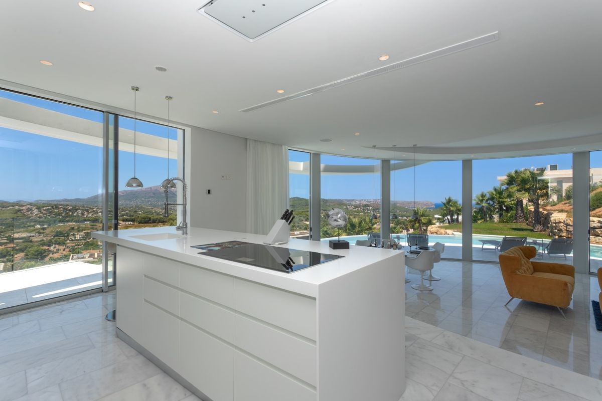 Cuisine moderne à plan ouvert avec îlot blanc épuré, sol en marbre et baies vitrées offrant une vue panoramique sur les collines et la piscine.