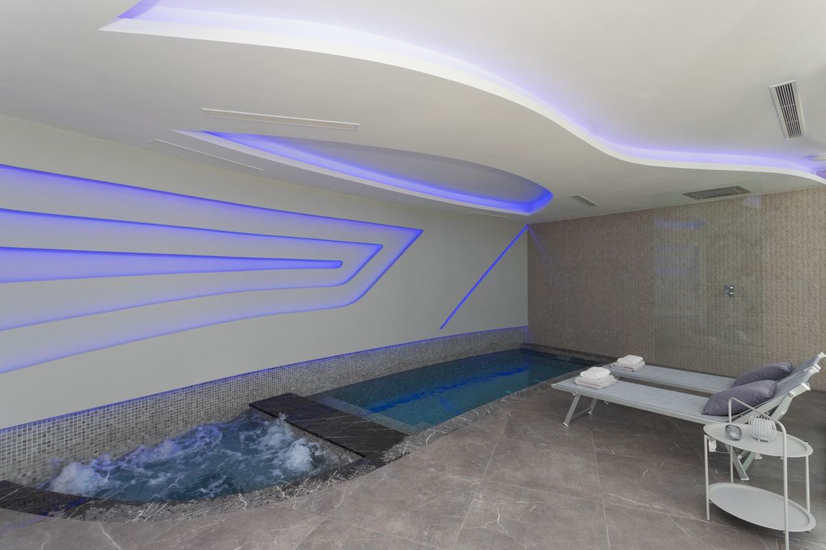 Espace bien-être intérieur comprenant un bassin avec jacuzzi, éclairage LED futuriste, revêtement de sol en pierre grise et chaises longues de relaxation.