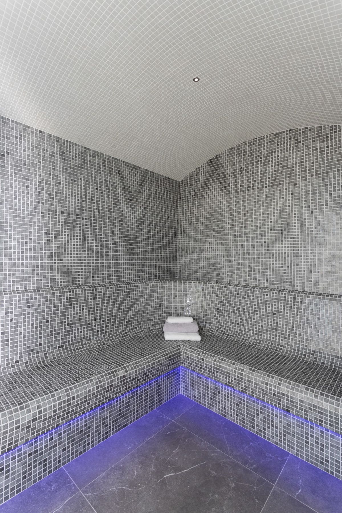 Hammam moderne avec parois en mosaïque grise, bancs ergonomiques éclairés par des LED bleues et plafond voûté en tesselles blanches.