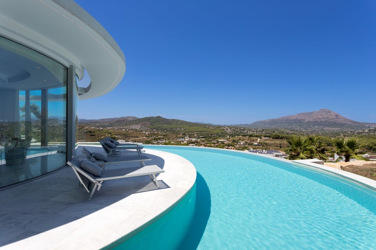 Piscine à débordement incurvée avec eau turquoise et terrasse en pierre blanche équipée de transats gris, offrant une vue panoramique sur la vallée.
