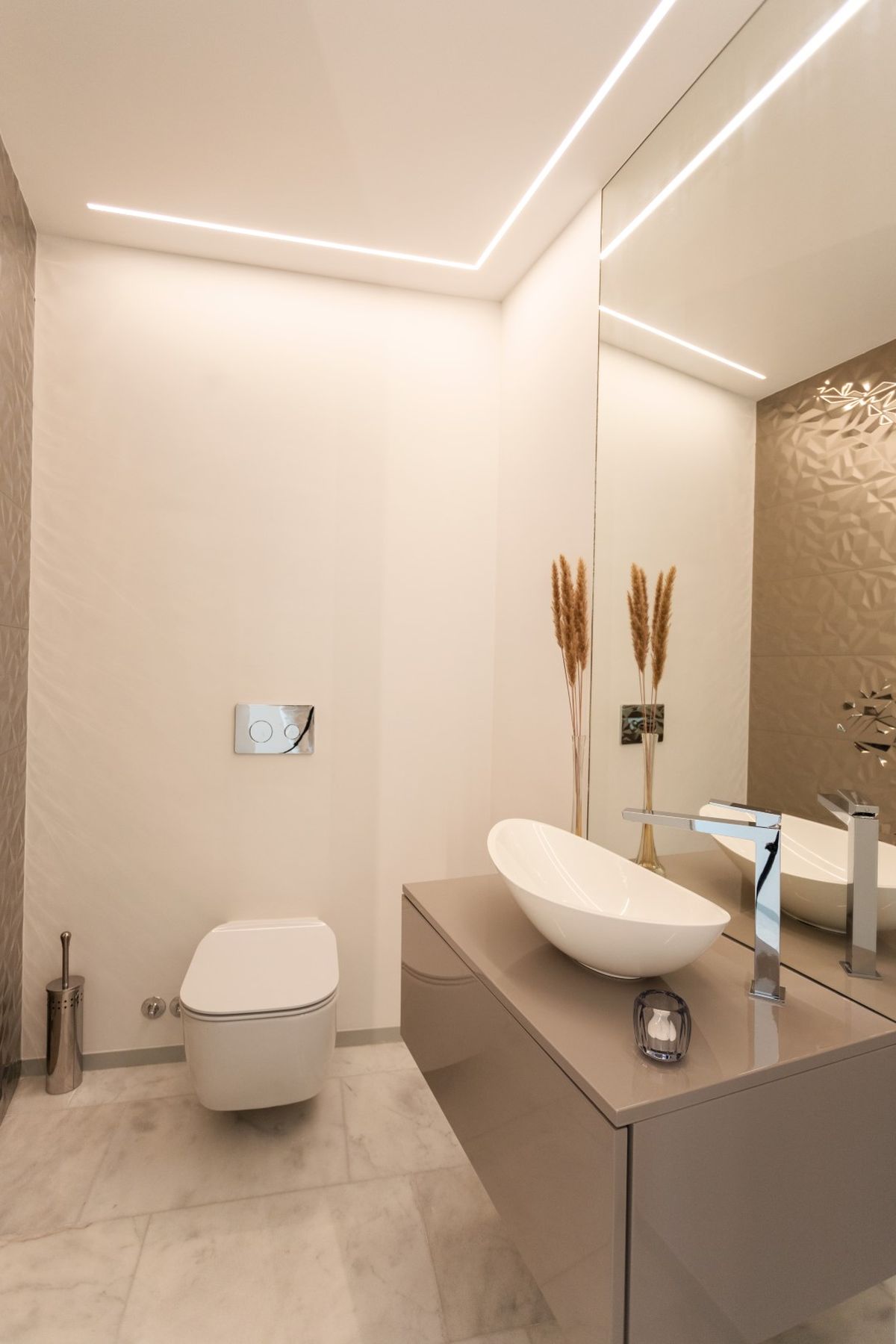 Salle d'eau moderne avec vasque à poser ovale, meuble suspendu taupe et WC suspendu. L'éclairage LED linéaire souligne le mur à texture géométrique.