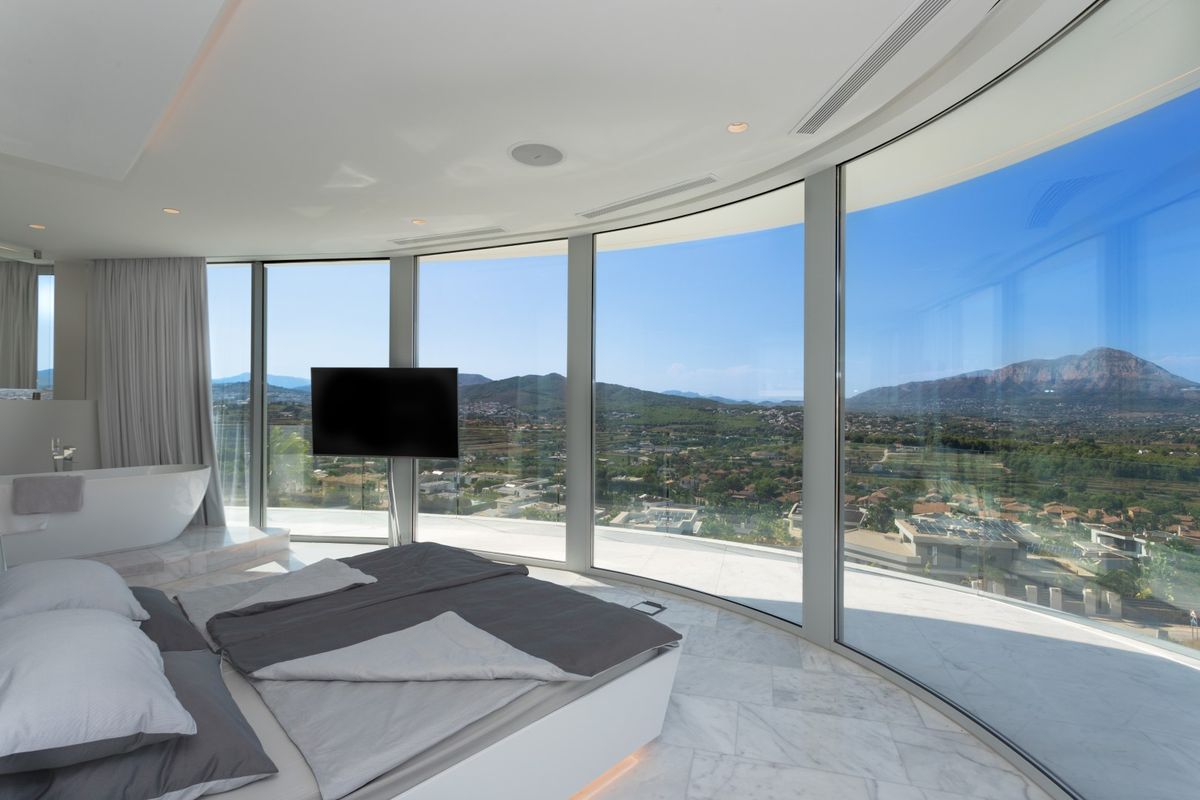 Suite parentale avec paroi vitrée courbe offrant une vue panoramique sur la vallée, sol en marbre blanc et baignoire îlot intégrée.