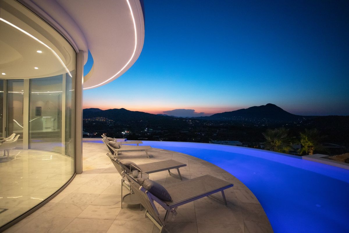 Terrasse avec piscine à débordement et éclairage LED bleu au crépuscule, architecture en verre courbé et vue panoramique sur les montagnes environnantes.