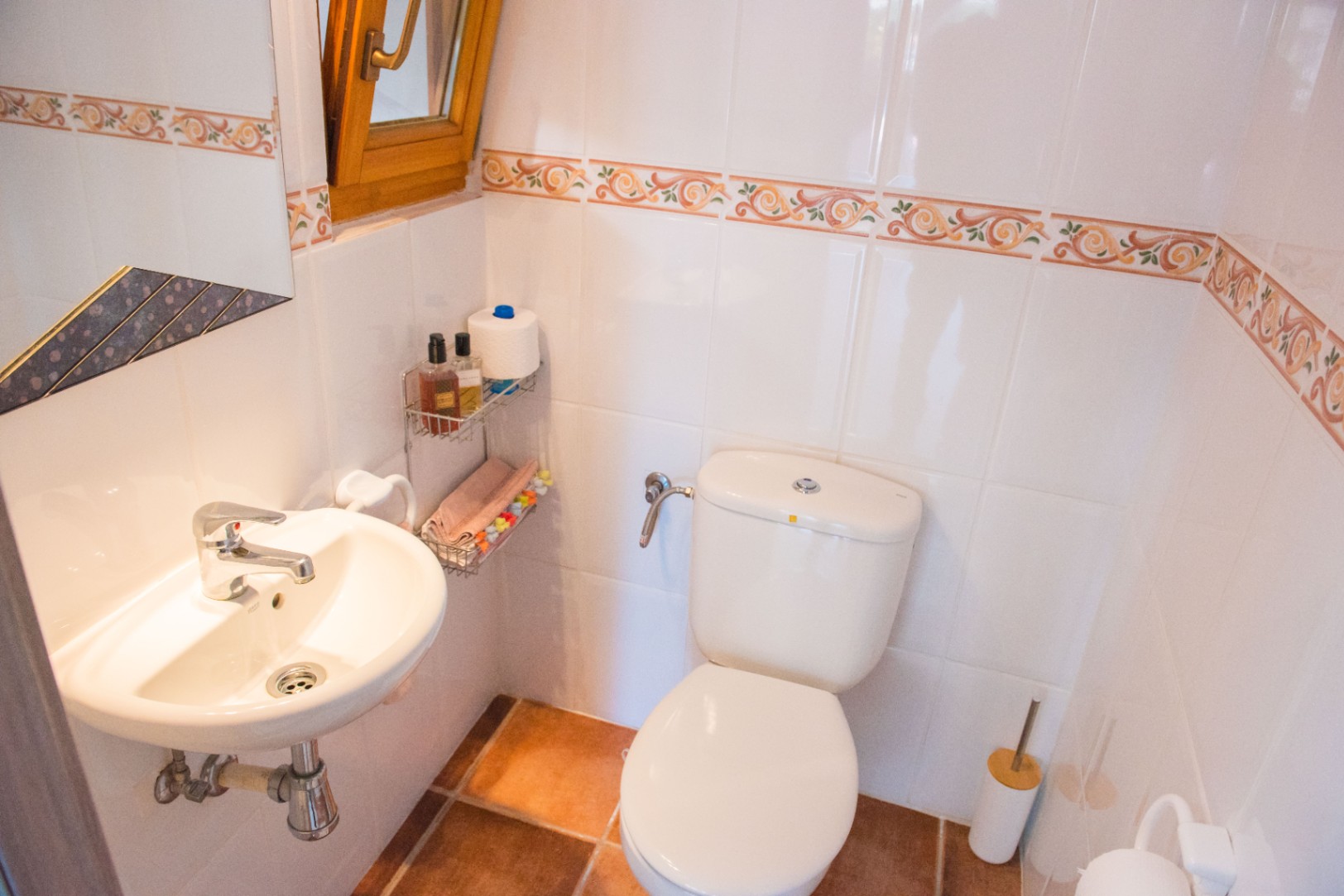 Cabinet de toilette compact avec carrelage mural blanc à frise décorative, sol en terre cuite, lavabo mural et fenêtre à cadre en bois pour la ventilation.