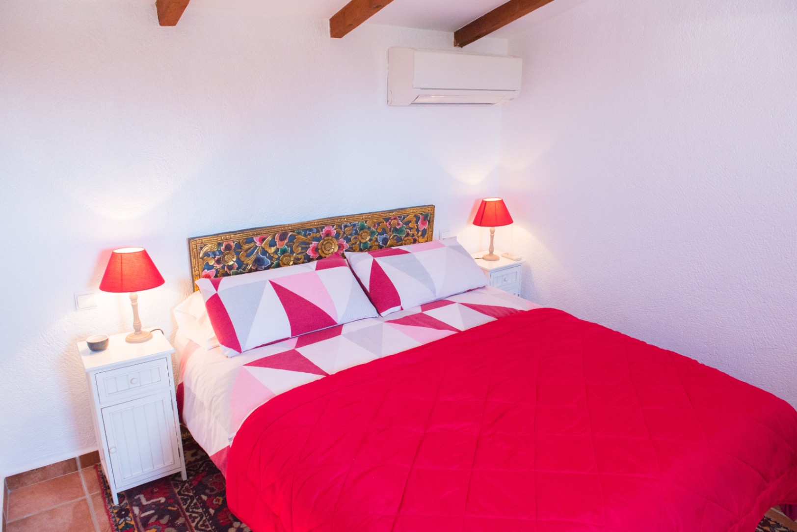 Chambre avec poutres apparentes, tête de lit sculptée colorée et tables de chevet blanches. Équipée de la climatisation et de sols en terre cuite.