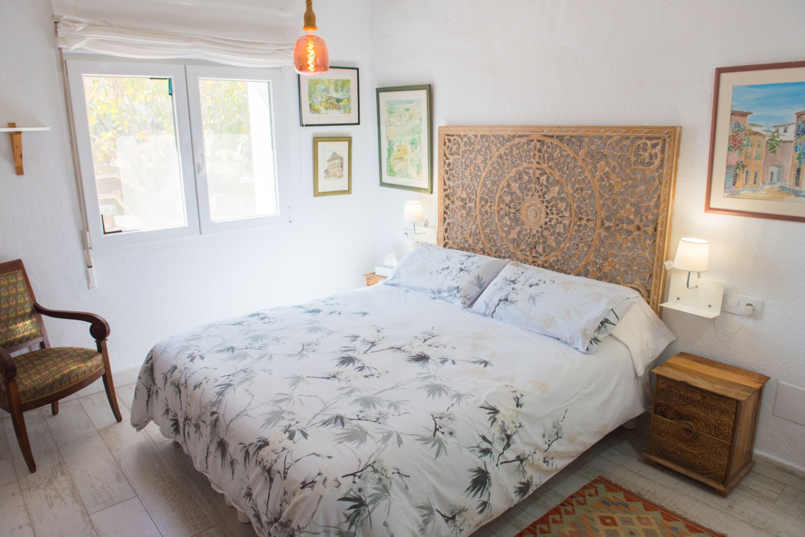 Chambre avec tête de lit en bois sculpté, literie à motifs floraux et fauteuil vintage. La pièce dispose de fenêtres en PVC blanc et d'un éclairage suspendu.