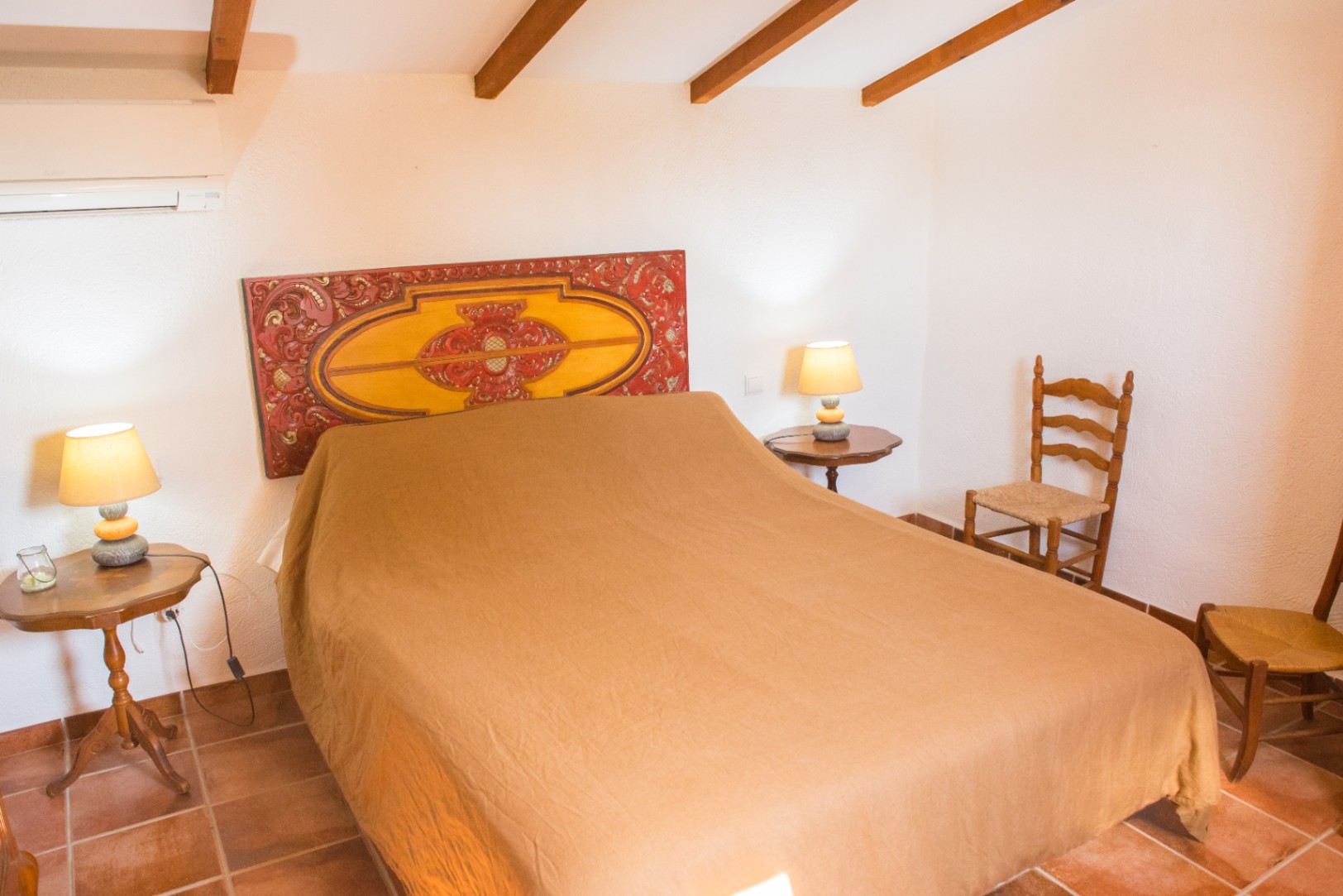 Chambre avec tête de lit en bois sculpté, poutres de plafond apparentes, sol en terre cuite et mobilier rustique avec chaises en osier.