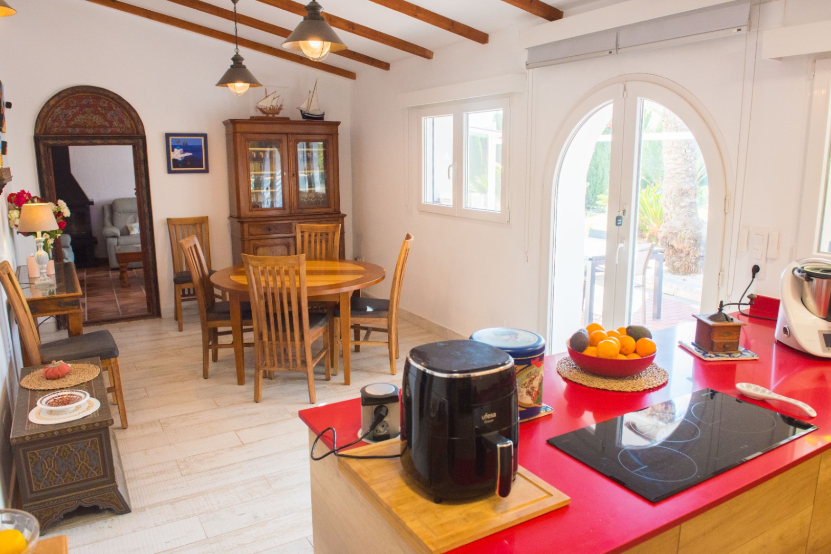 Cuisine avec îlot central rouge, coin repas en bois, poutres apparentes et porte vitrée en arche ouvrant sur l'extérieur.