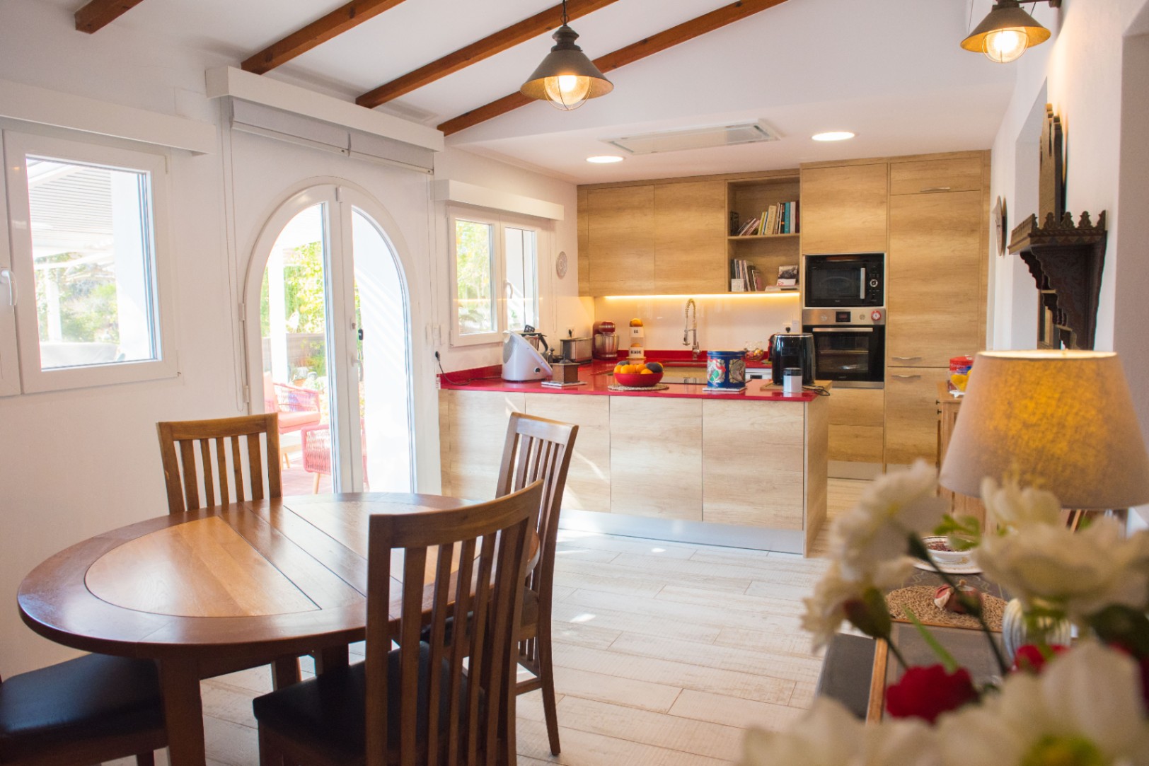 Cuisine moderne avec meubles en bois clair, plan de travail rouge vif et poutres apparentes au plafond, complétée par un coin repas avec table circulaire.