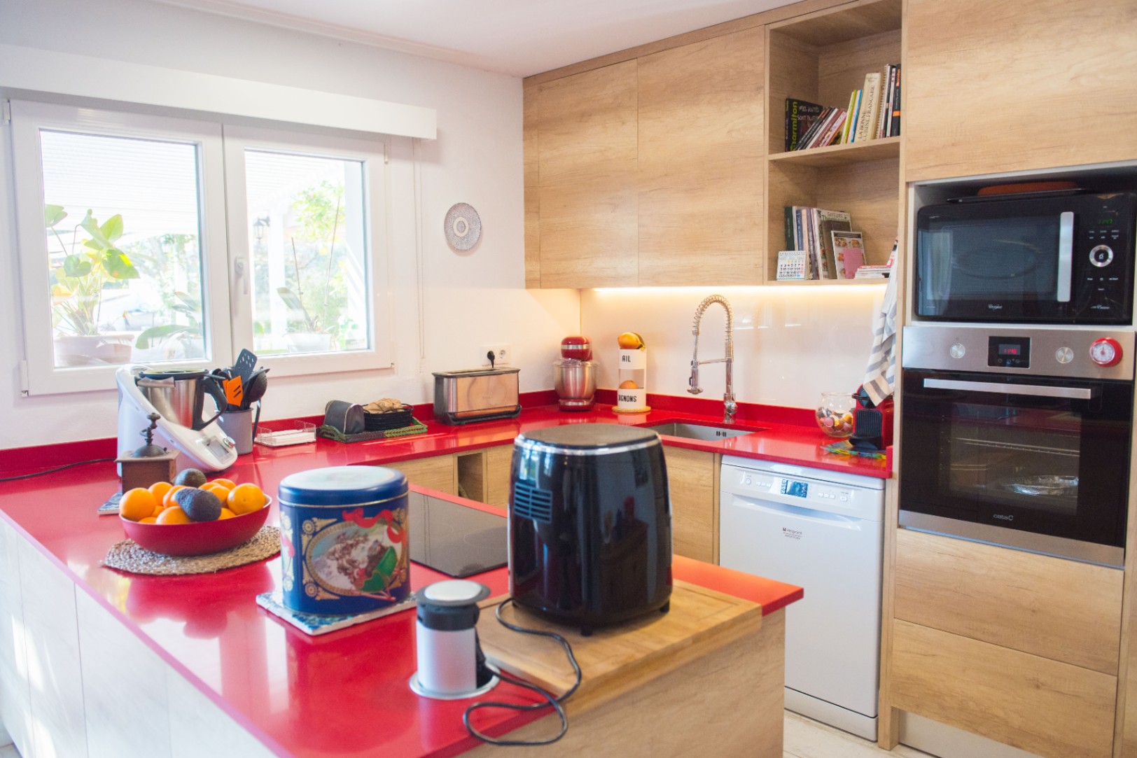 Cuisine moderne avec plan de travail rouge vif, meubles en bois clair et appareils intégrés, comprenant un four, un micro-ondes et un lave-vaisselle.