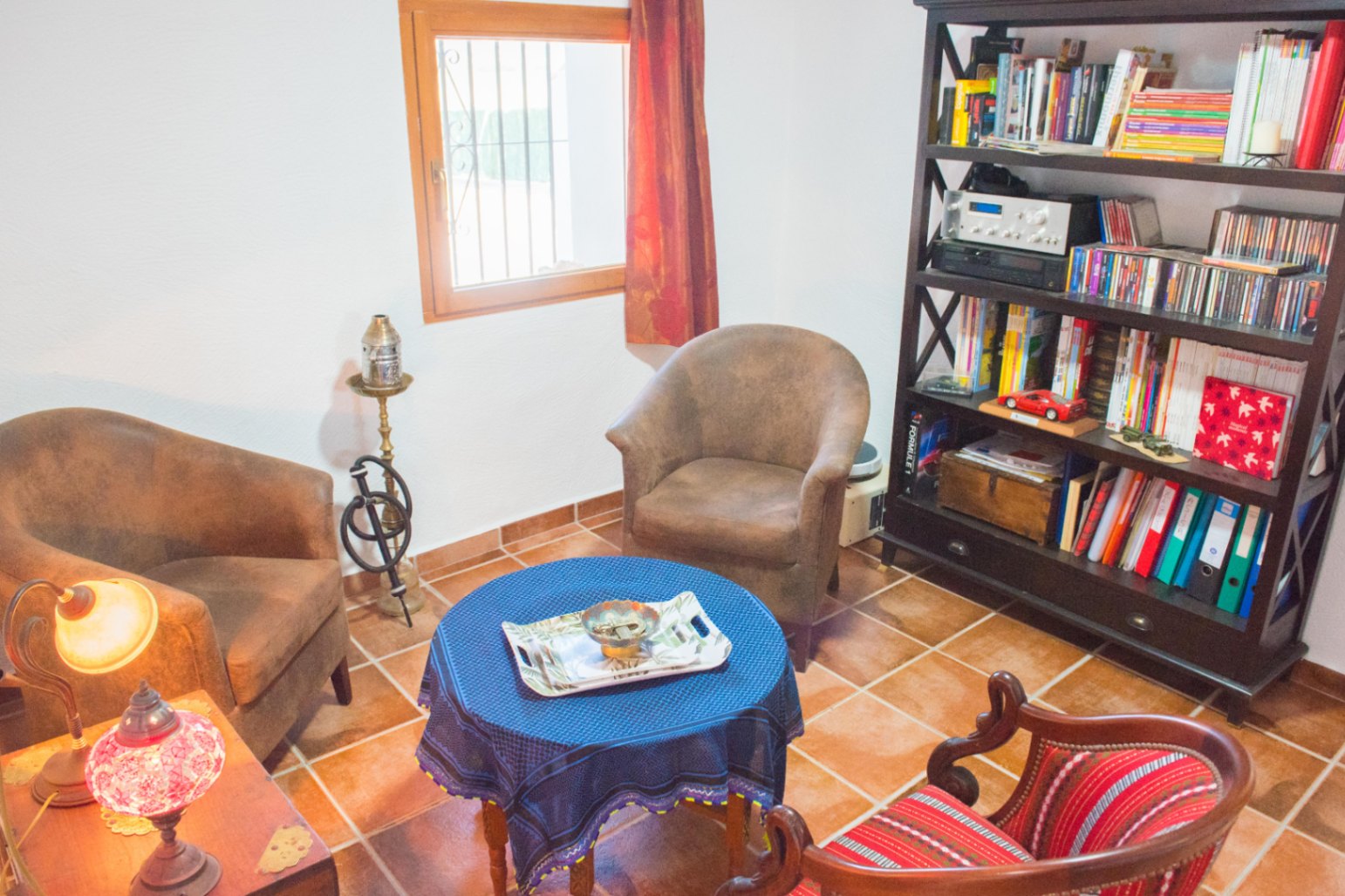 Espace de lecture avec sol en terre cuite, bibliothèque en bois sombre et fauteuils entourant une table ronde recouverte d'une nappe bleue.
