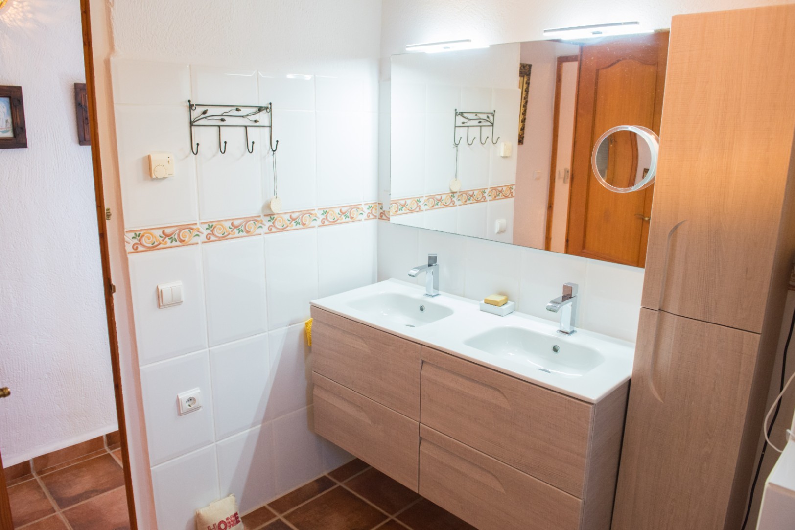 Meuble vasque double suspendu en bois clair avec éviers intégrés blancs, robinetterie chromée et grand miroir équipé de luminaires LED et d'un miroir grossissant.