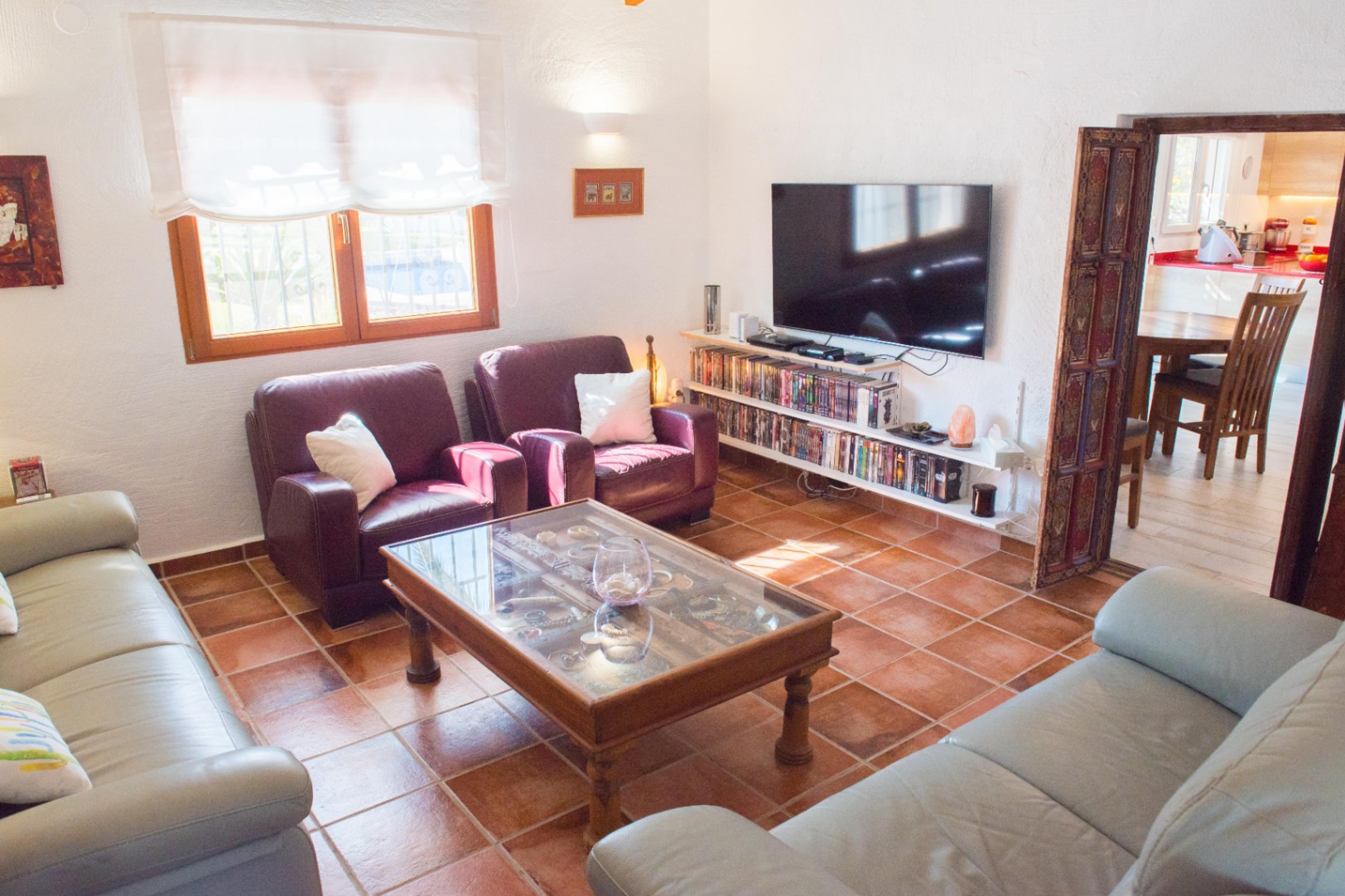 Salon avec sol en terre cuite, canapés en cuir et table basse vitrée. Comprend des étagères multimédia sous le téléviseur et un encadrement de porte en bois sculpté.