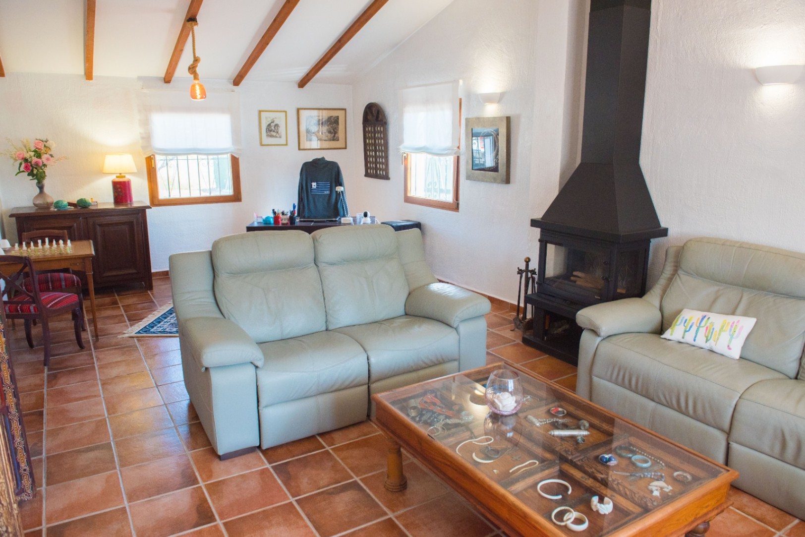 Salon avec sol en terre cuite, cheminée en métal noir et plafond mansardé avec poutres en bois apparentes. Comprend des canapés en cuir et une table basse vitrée.