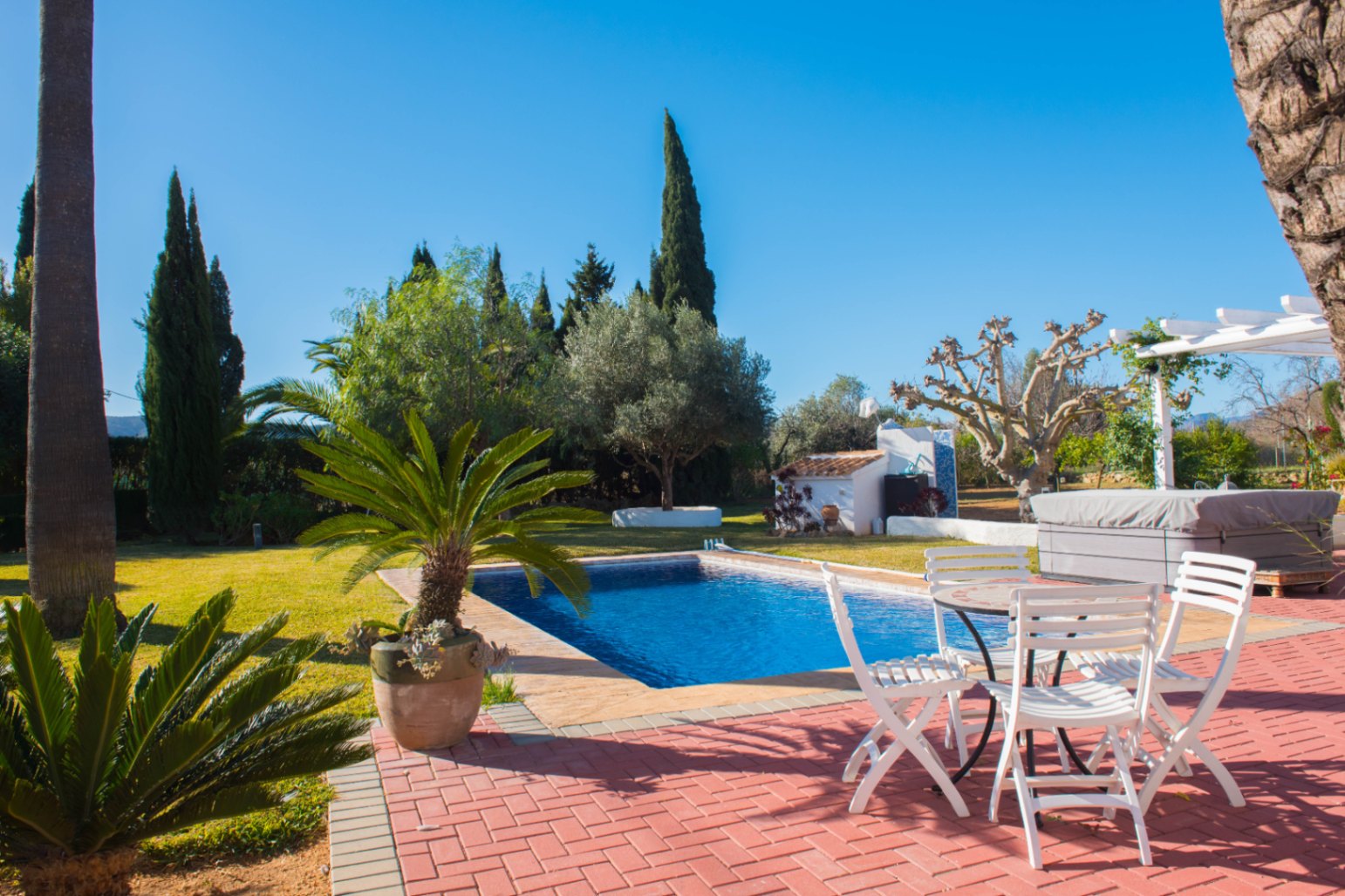 Terrasse extérieure en briques rouges comprenant une piscine rectangulaire, du mobilier de jardin blanc et un jardin paysager avec des cyprès.