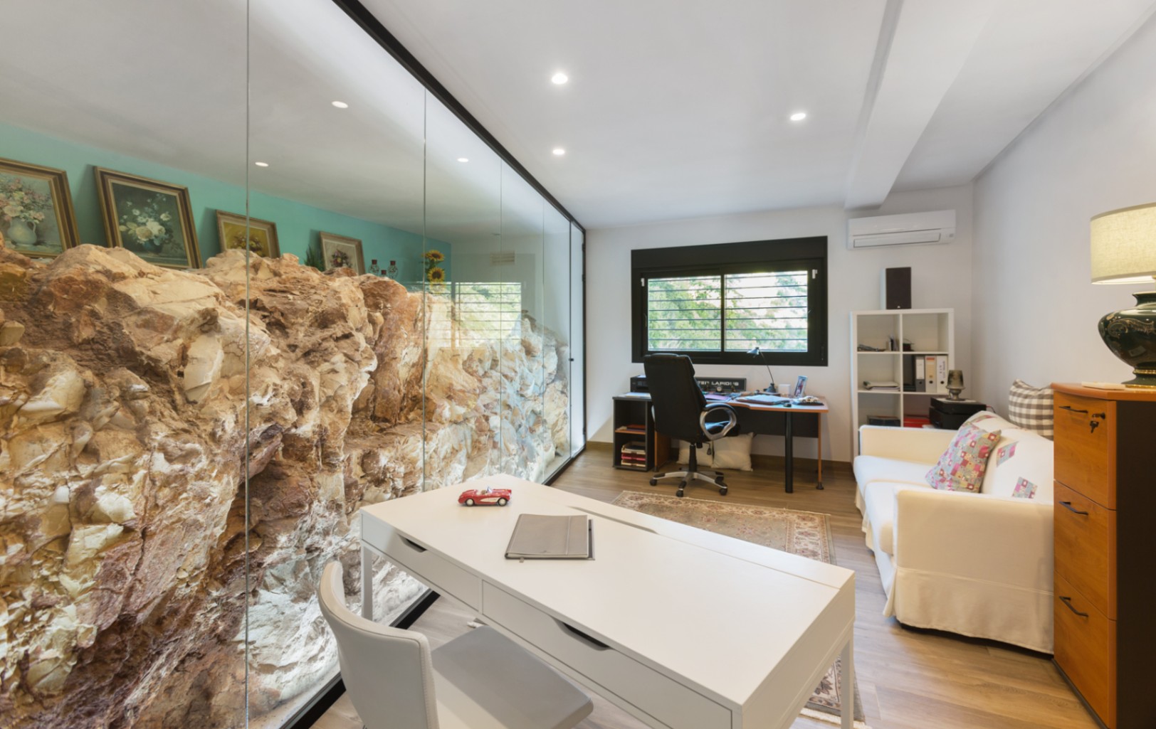 Bureau moderne doté d'une paroi vitrée intégrant une formation rocheuse naturelle. Comprend un bureau blanc, un canapé et un éclairage au plafond encastré.