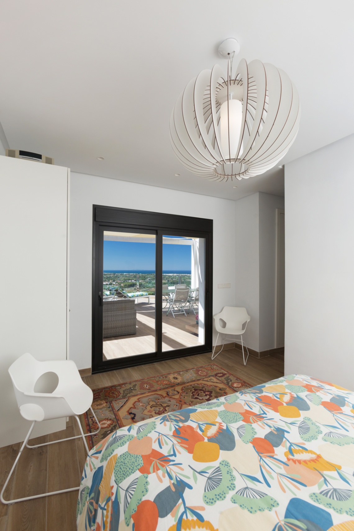 Chambre lumineuse avec accès direct à la terrasse par des baies vitrées noires, luminaire design et vue panoramique sur la mer.