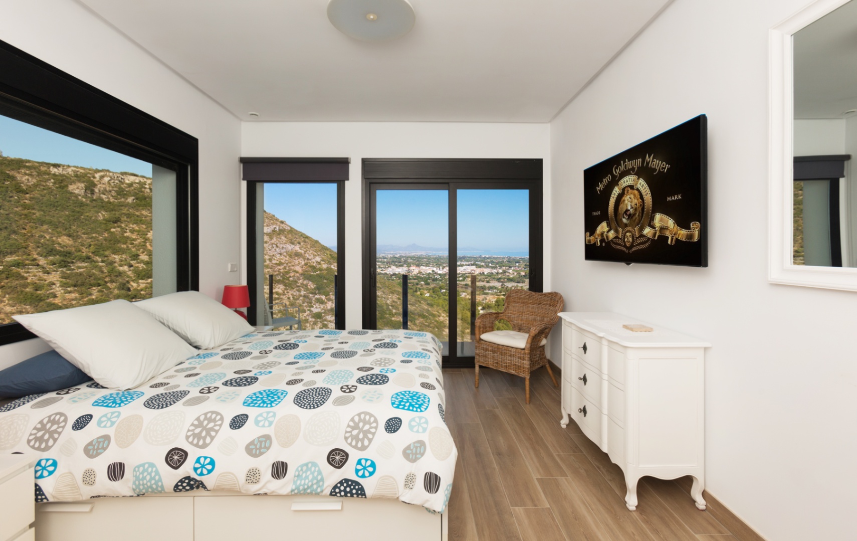Chambre lumineuse avec lit design, parquet et grandes fenêtres noires encadrant des vues panoramiques sur la montagne et la côte lointaine. Comprend une commode blanche et une télévision. Chambre lumineuse avec lit design, parquet et grandes fenêtres noires encadrant des vues panoramiques sur la montagne et la côte lointaine. Comprend une commode blanche et une télévision.