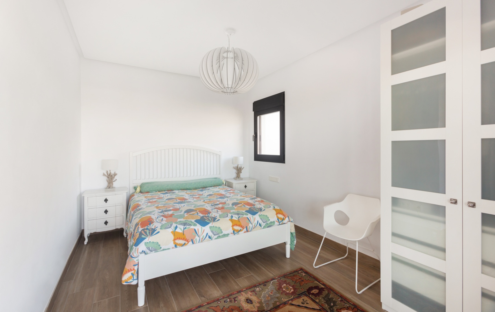 Chambre lumineuse avec lit en bois blanc, tables de chevet assorties et armoire à panneaux dépolis. Sol en grès cérame aspect bois et suspension design moderne.