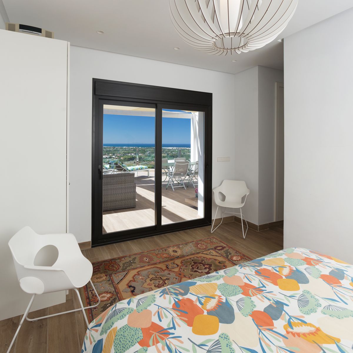 Chambre moderne avec baie vitrée coulissante ouvrant sur une terrasse meublée offrant une vue sur la mer. Comprend un luminaire suspendu sculptural et des chaises design blanches.