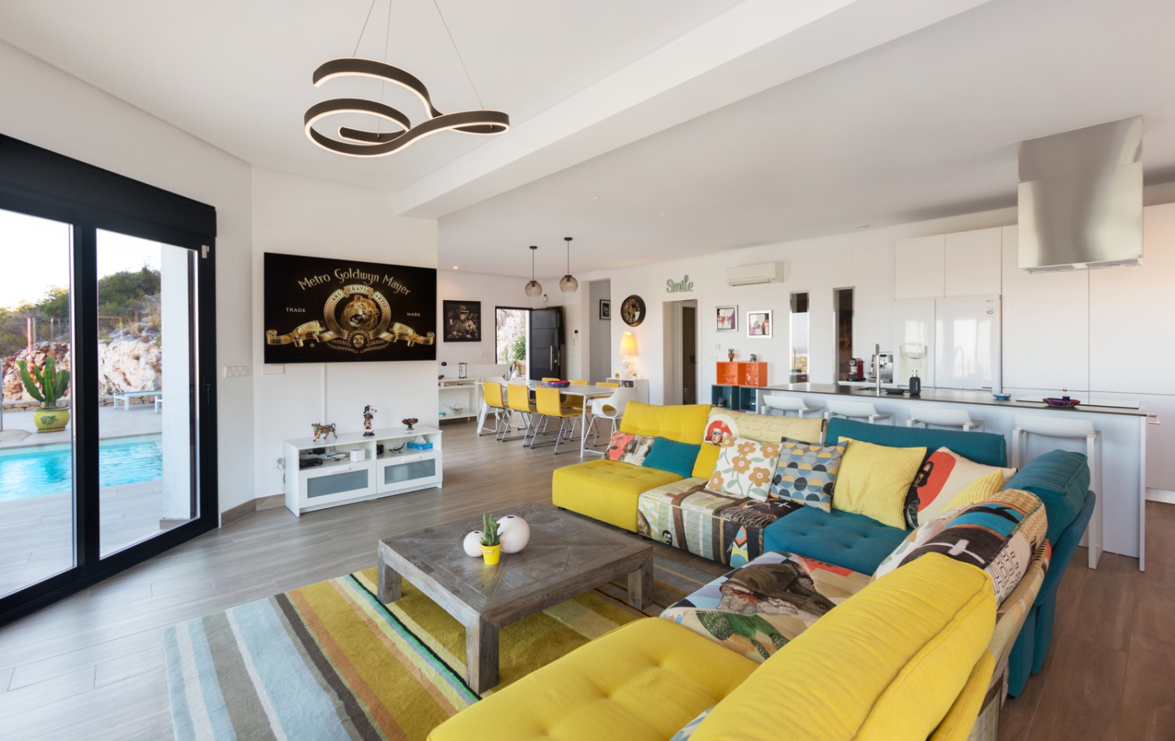 Espace de vie ouvert comprenant un canapé modulaire coloré, une table basse en bois et un accès à la terrasse avec piscine par de grandes baies vitrées.