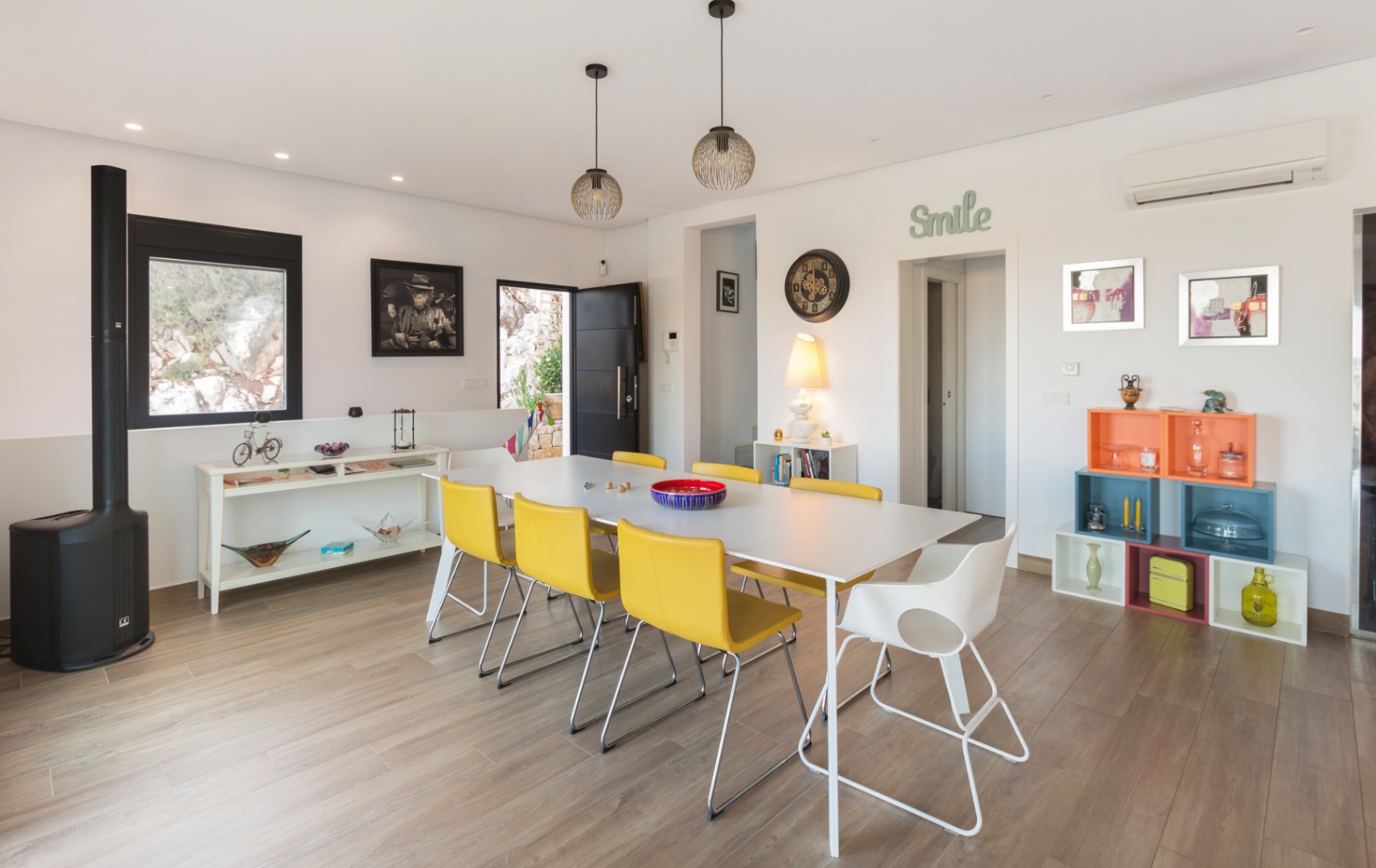 Espace salle à manger moderne avec table blanche, chaises jaunes, sol imitation bois, poêle à bois noir et étagères modulaires aux couleurs vives.