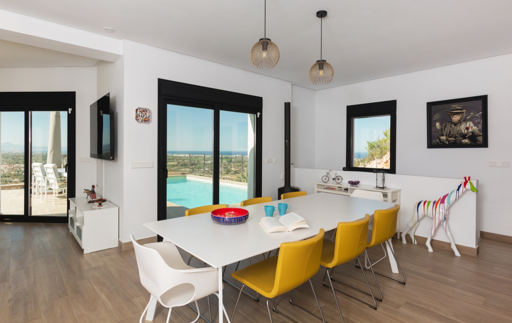 Salle à manger moderne avec table blanche et chaises jaunes, sol effet bois et accès terrasse avec piscine offrant une vue dégagée sur la côte et le paysage.