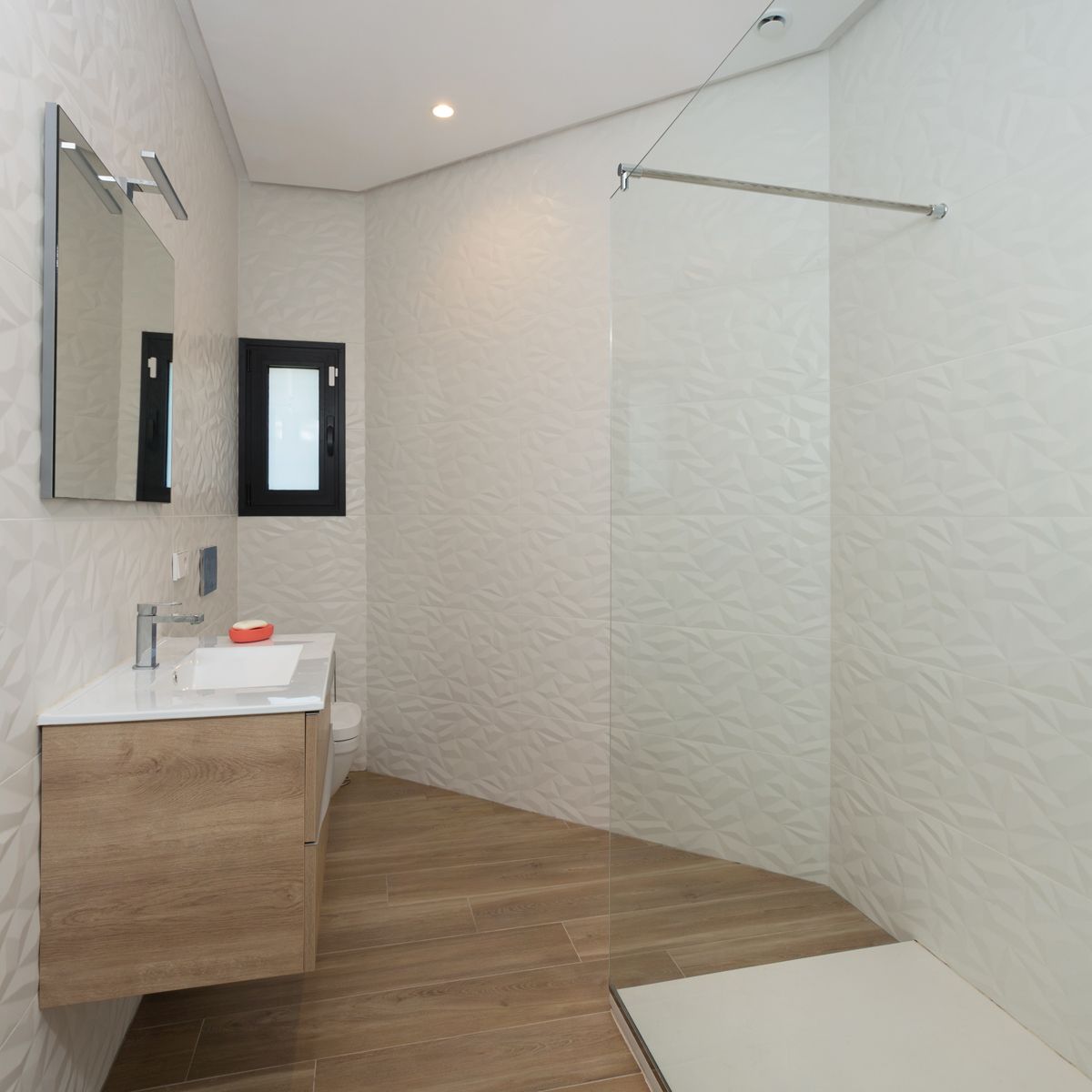 Salle de bain moderne avec meuble-lavabo suspendu en bois, vasque blanche, miroir éclairé et douche à l'italienne vitrée. Carrelage mural blanc texturé géométrique et sol imitation bois. Salle de bain moderne avec meuble-lavabo suspendu en bois, vasque blanche, miroir éclairé et douche à l'italienne vitrée. Carrelage mural blanc texturé géométrique et sol imitation bois.