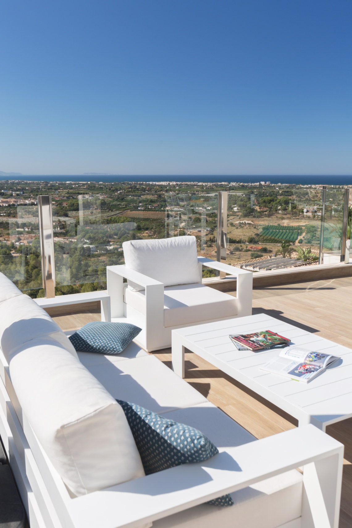 Terrasse avec mobilier de salon blanc moderne et garde-corps en verre, offrant une vue panoramique sur le paysage côtier et la mer Méditerranée.