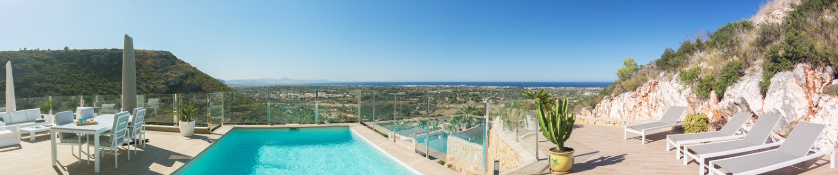 Terrasse surélevée avec piscine, garde-corps en verre et mobilier blanc, offrant une vue panoramique sur la vallée et la côte méditerranéenne.