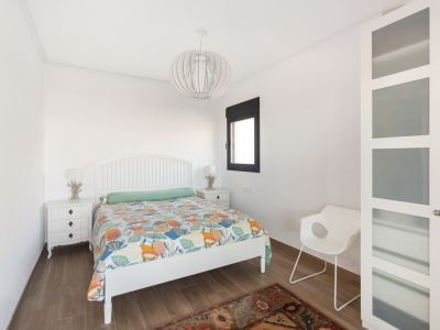 Chambre avec tête de lit à lattes blanche, couette colorée et carrelage imitation bois. Comprend des tables de chevet blanches avec lampes décoratives, une fenêtre et une armoire à panneaux de verre dépoli.