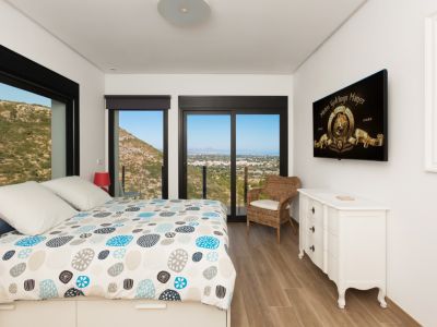 Chambre lumineuse avec lit design, parquet et grandes fenêtres noires encadrant des vues panoramiques sur la montagne et la côte lointaine. Comprend une commode blanche et une télévision.