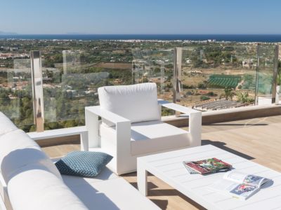 Espace salon extérieur moderne avec mobilier blanc sur une terrasse en bois, doté d'une balustrade en verre offrant une vue panoramique sur la côte et la mer Méditerranée.