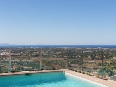 Piscine à débordement avec eau bleue claire et balustrade en verre, offrant des vues panoramiques sur le paysage côtier et la mer Méditerranée sous un ciel clair.
