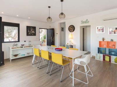 Salle à manger lumineuse avec une longue table blanche, six chaises jaunes et une blanche, éclairée par des suspensions modernes. Sol carrelé imitation bois et décor éclectique.