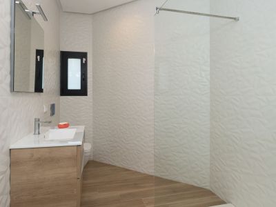 Salle de bain moderne avec meuble-lavabo suspendu en bois, vasque blanche, miroir éclairé et douche à l'italienne vitrée. Carrelage mural blanc texturé géométrique et sol imitation bois.