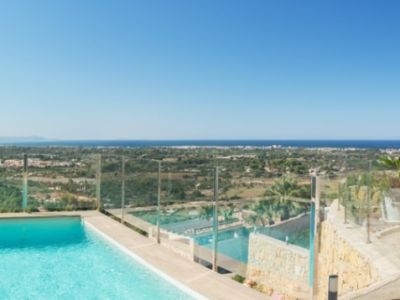 Vaste terrasse extérieure avec piscine rectangulaire, mobilier blanc moderne et chaises longues. Vues panoramiques sur la côte, la mer et les collines verdoyantes sous un ciel clair.