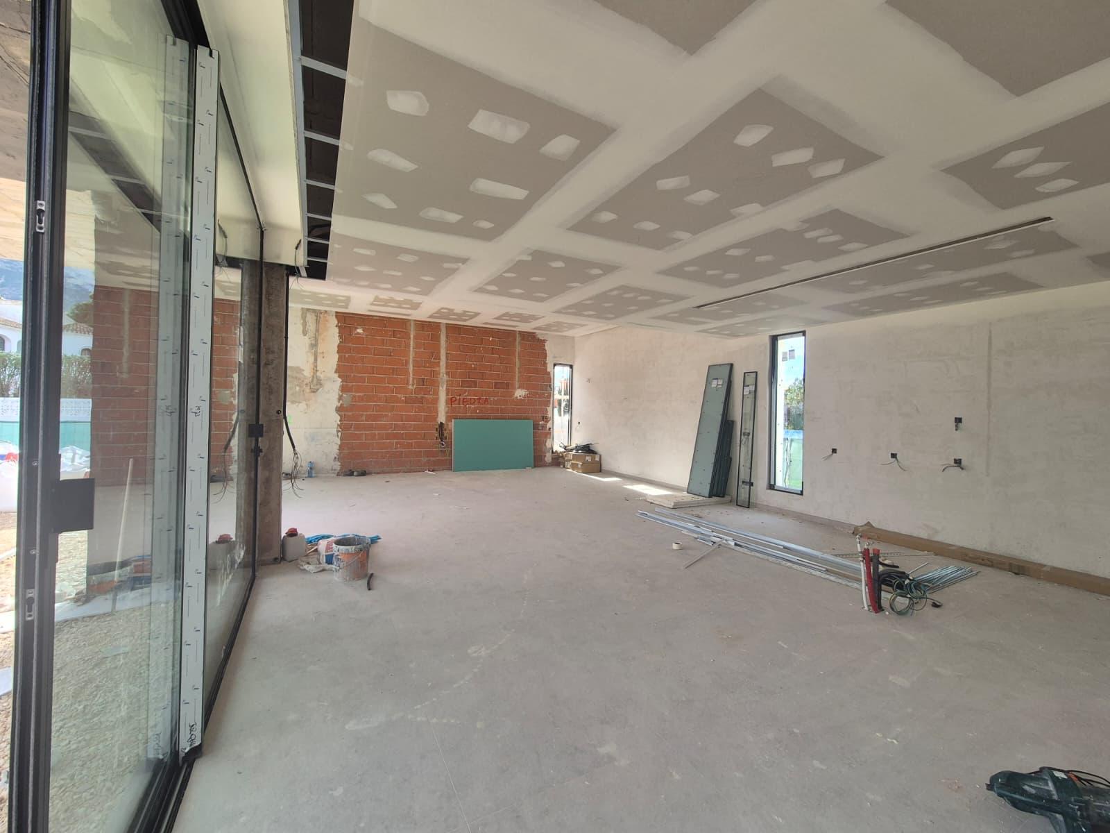 Grand salon en cours de construction avec plafond en plaques de plâtre, mur en briques apparentes et larges baies vitrées coulissantes.