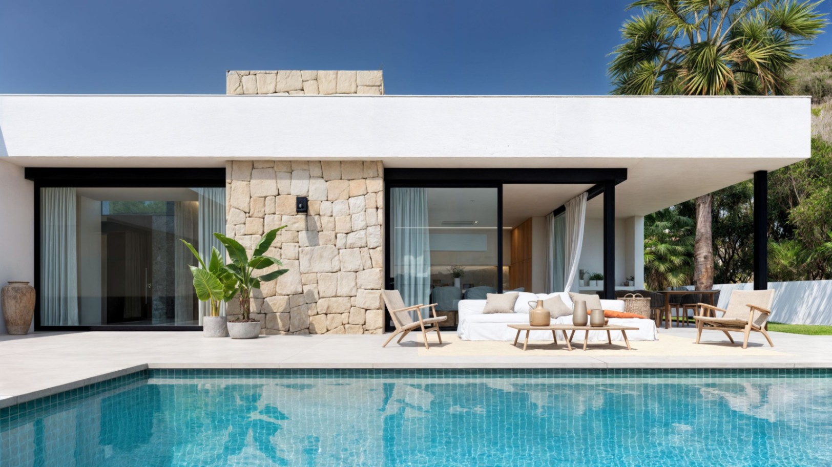 Façade de villa moderne avec piscine turquoise, murs en pierre apparente et salon extérieur aménagé sur une terrasse en dalles de grand format.