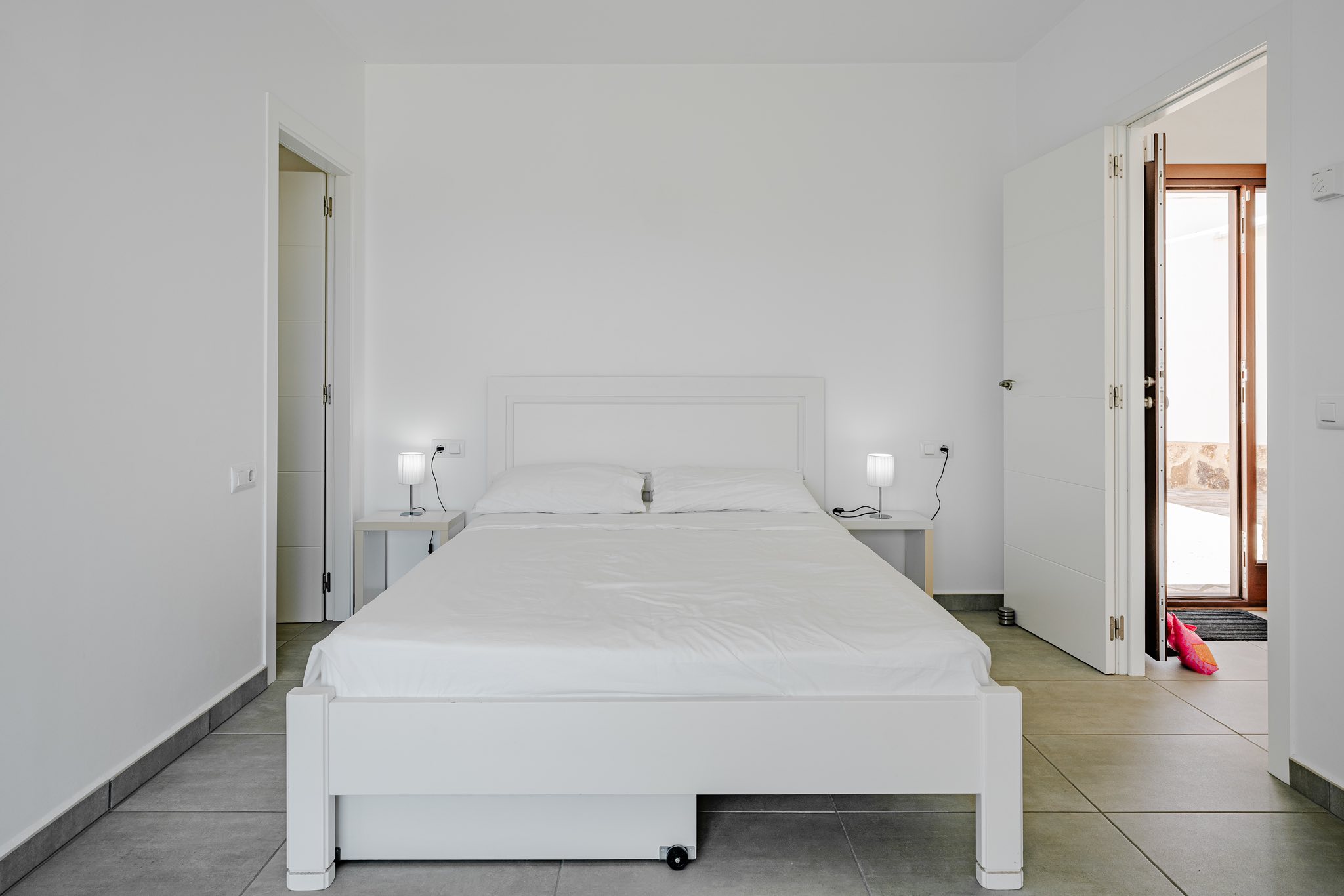 Chambre minimaliste avec lit en bois blanc, tables de chevet assorties et sol en carrelage gris. Les portes ouvertes donnent sur l'extérieur et les pièces adjacentes.