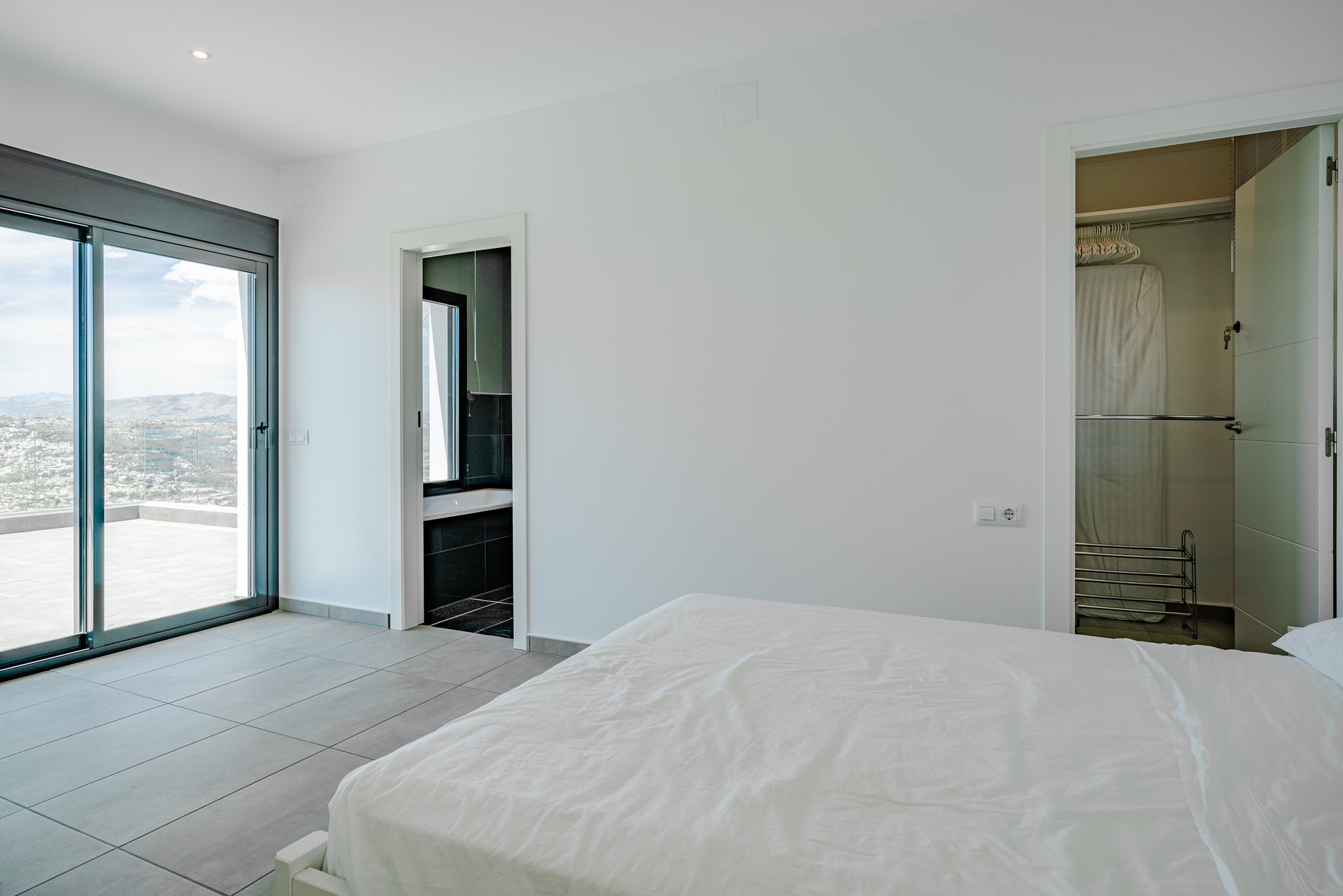 Chambre moderne avec porte vitrée coulissante ouvrant sur une terrasse, salle de bain attenante en carrelage sombre et dressing intégré.