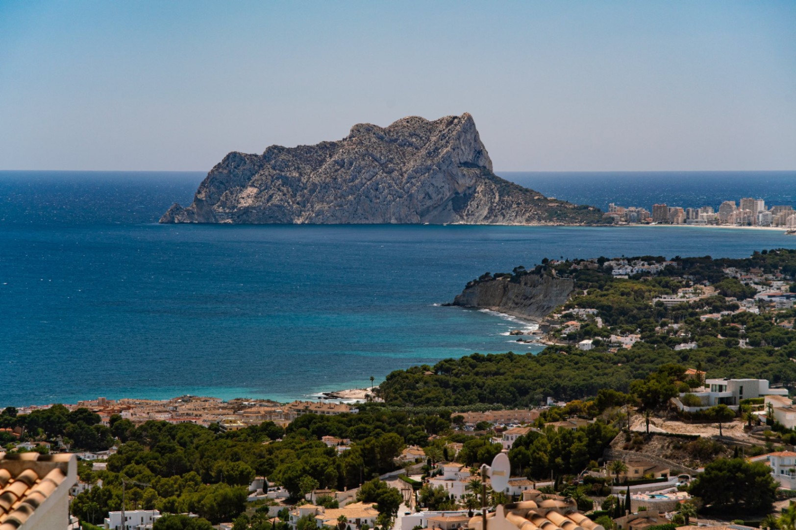 Vue panoramique surélevée de la côte méditerranéenne mettant en valeur le Peñón de Ifach, les eaux turquoise et un quartier résidentiel entouré de pins.