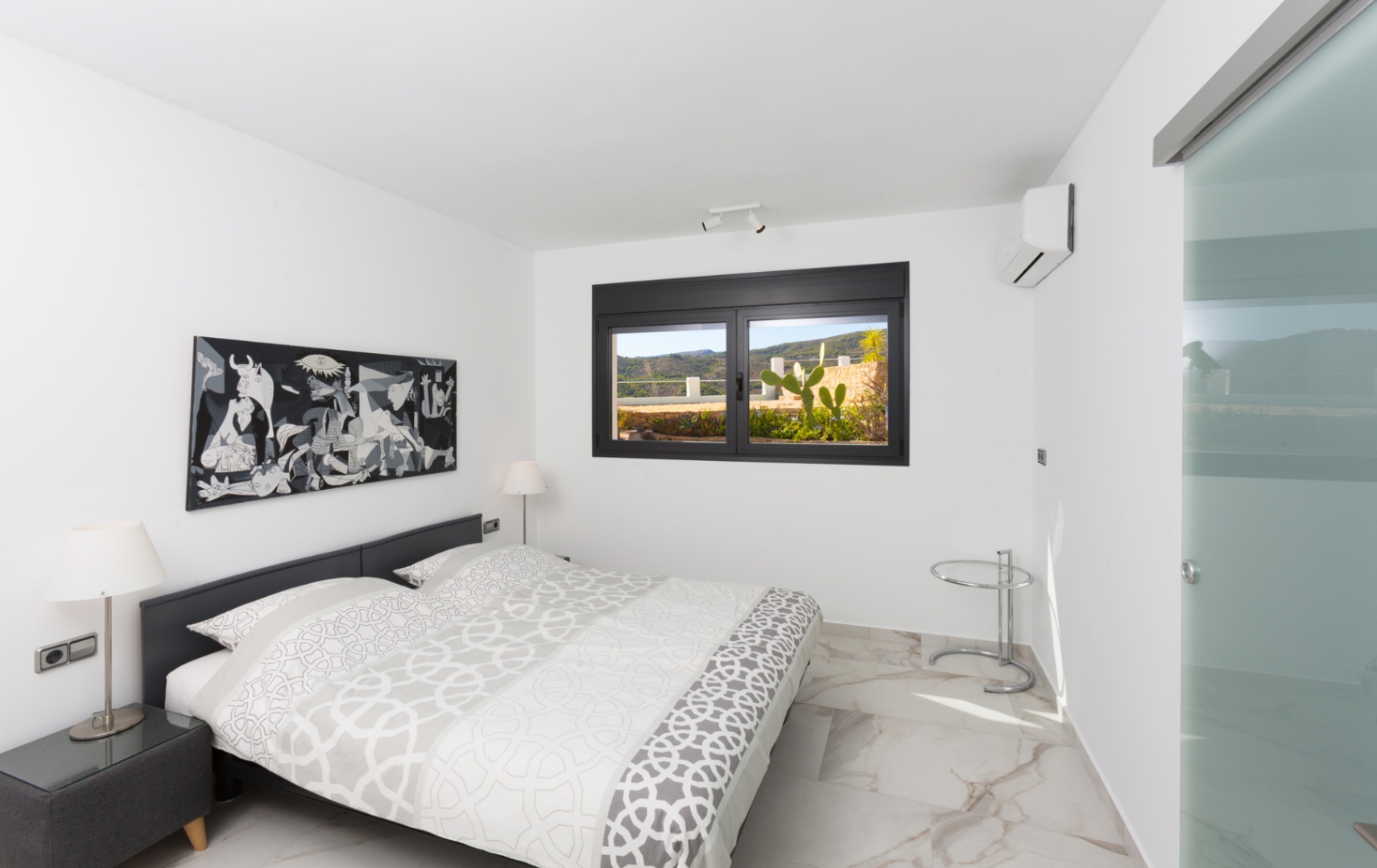 Chambre moderne avec sol en grès cérame effet marbre, œuvre d'art monochrome et fenêtre à cadre noir offrant une vue sur une terrasse avec cactus.