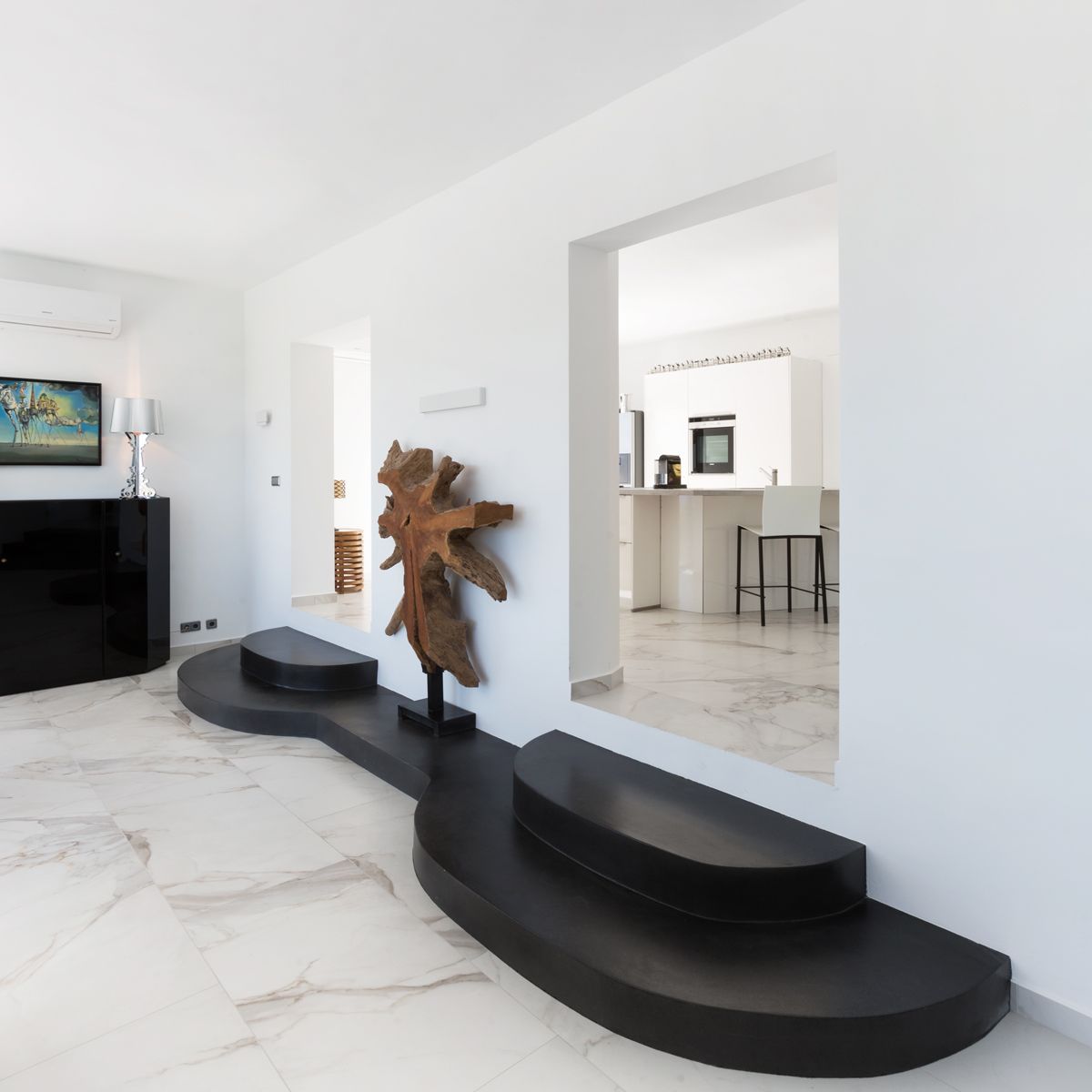 Espace de vie moderne avec sol en marbre blanc et plateforme noire incurvée. Une sculpture en bois naturel souligne l'ouverture vers une cuisine blanche épurée.