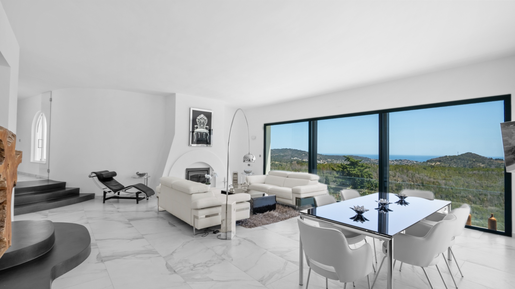 Espace de vie ouvert avec sol en marbre blanc, cheminée design et grandes baies vitrées offrant une vue panoramique sur les collines environnantes et la mer.