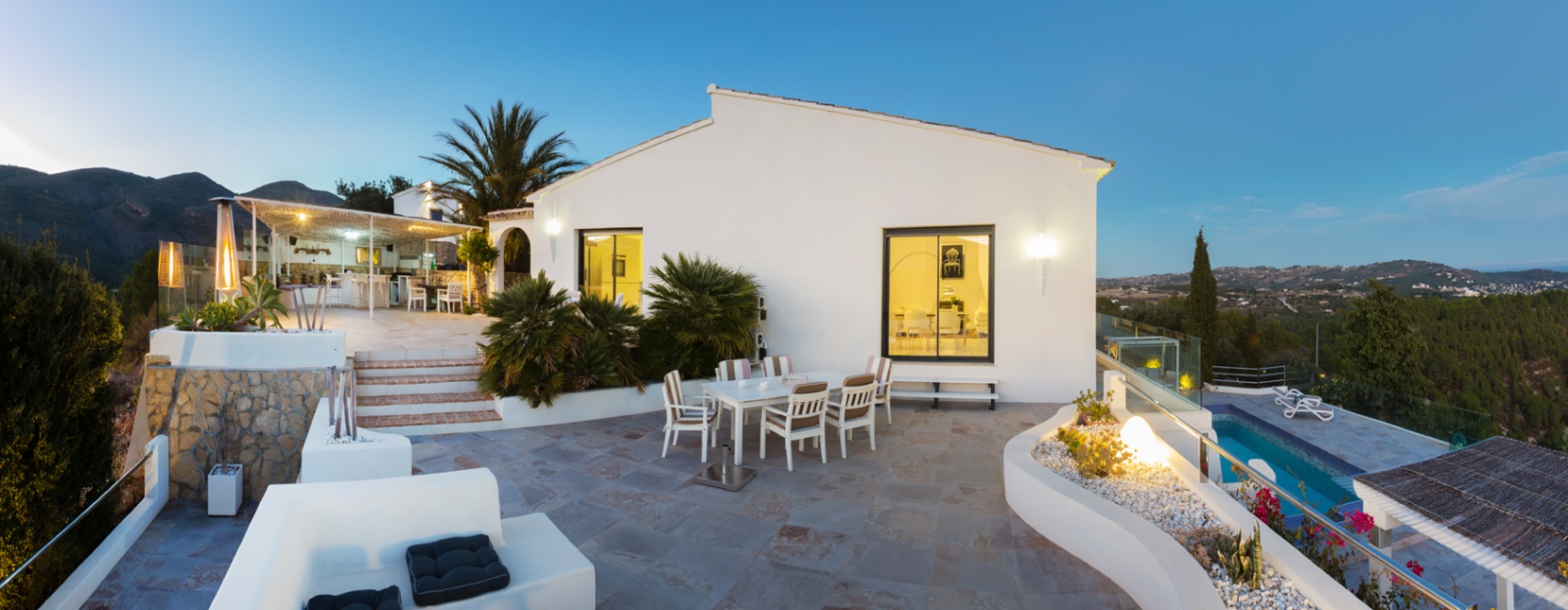 Grande terrasse en pierre au crépuscule avec coin repas, jardinières intégrées et vue panoramique sur la vallée, attenante à une villa méditerranéenne blanche.