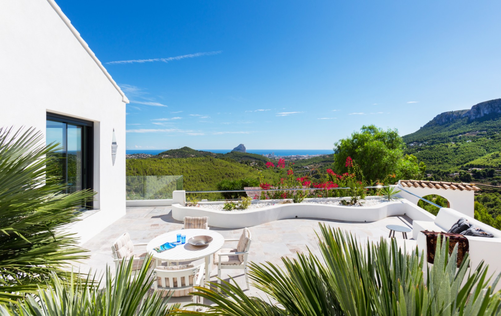 Grande terrasse pavée de pierre avec coin repas et jardinières blanches, offrant une vue panoramique sur la côte méditerranéenne et le Peñón de Ifach.