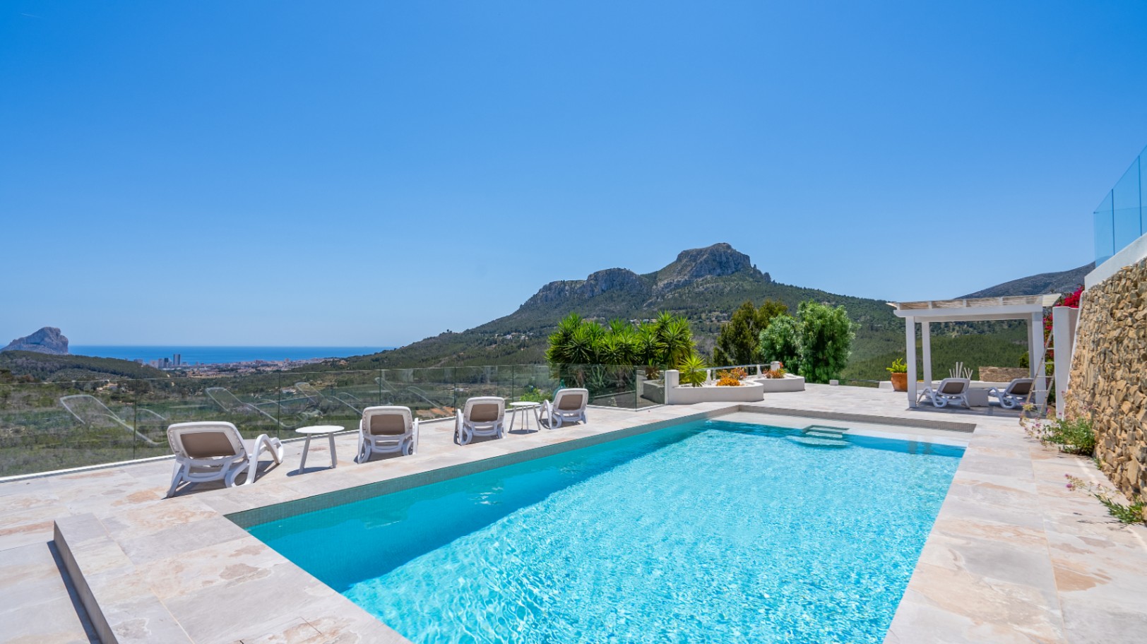 Piscine extérieure rectangulaire avec terrasse en pierre naturelle, garde-corps en verre et vue panoramique sur la côte et les montagnes.